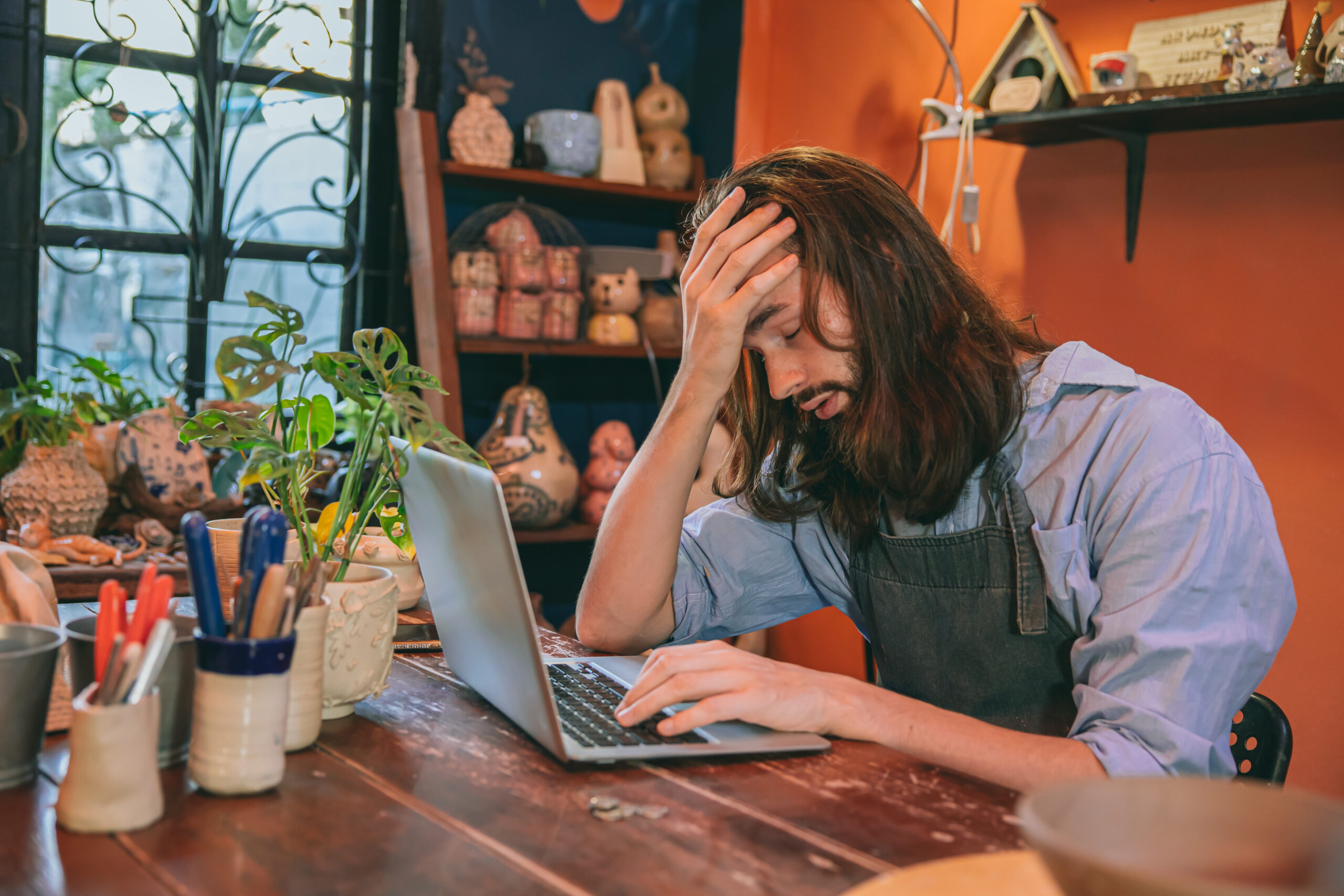 Ein Mann mit langen Haaren sitzt in seinem Atelier vor seinem Laptop und schaut verzweifelt mit der Hand vor dem Kopf auf die Tastatur